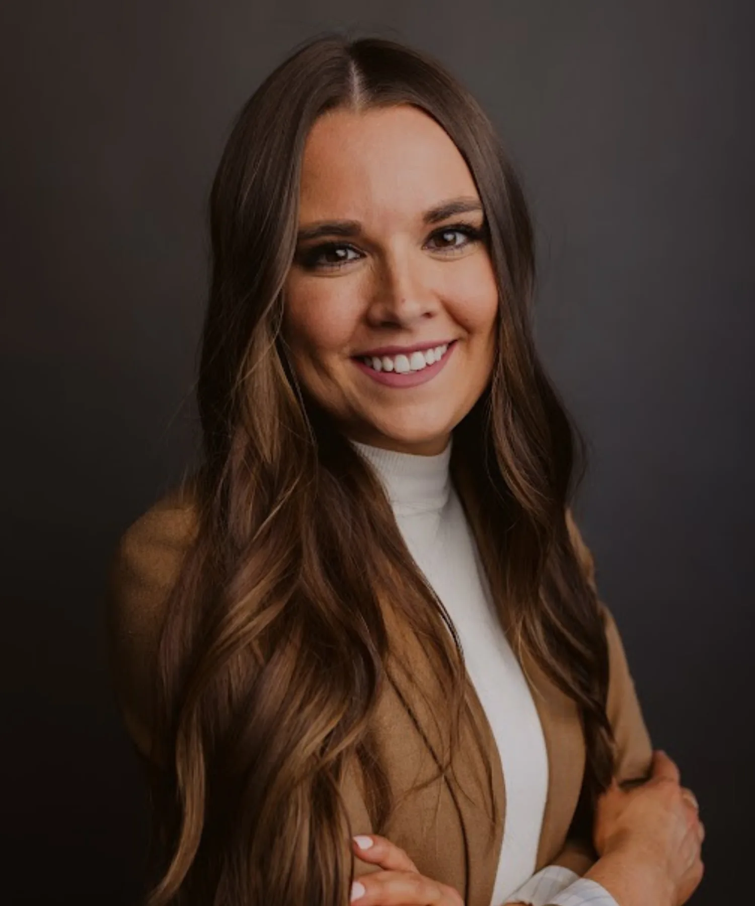 A woman with long, wavy brown hair smiling at the camera. She is wearing a tan blazer over a white top and has light makeup. The background is dark and neutral.