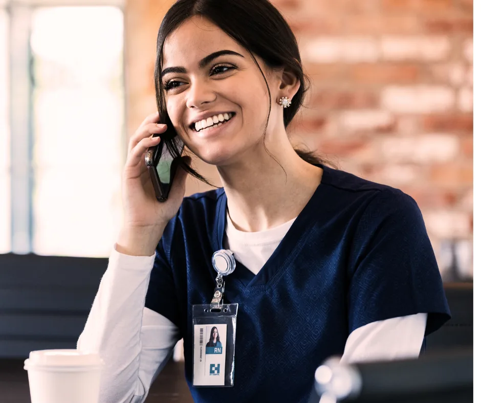 A smiling nurse with dark hair, wearing a navy blue uniform and a white undershirt, talking on the phone in a medical setting.
