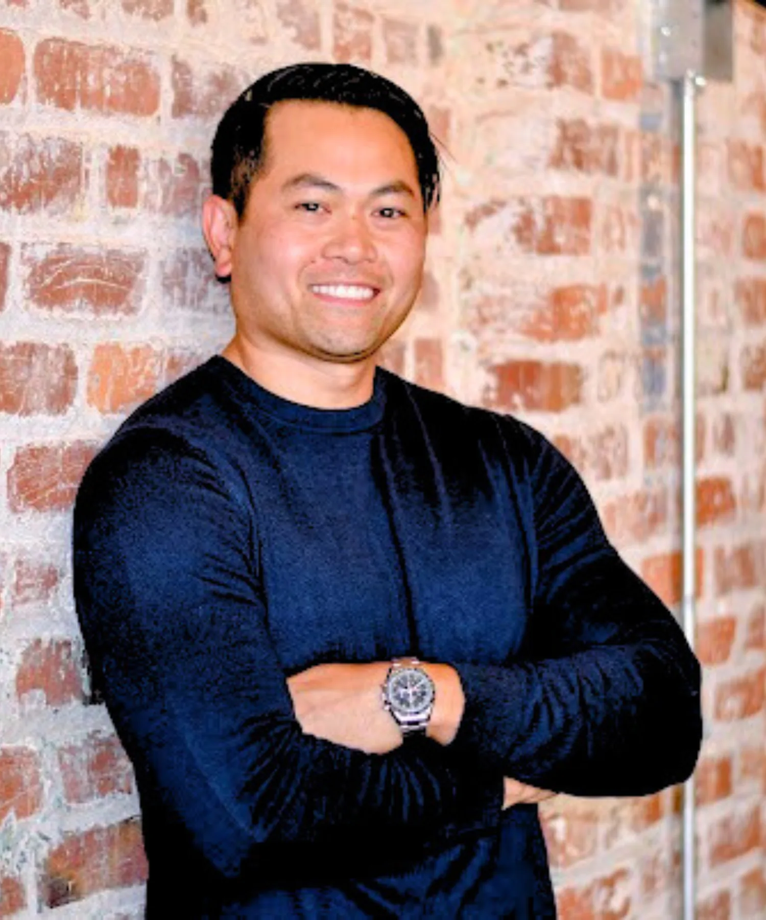 A smiling man with black hair standing with crossed arms in front of a brick wall.