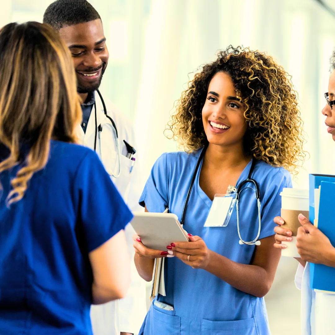 Group of diverse healthcare professionals, including nurses and doctors, engaged in a discussion in a medical setting. One woman in blue scrubs holds a tablet and smiles while speaking to colleagues.