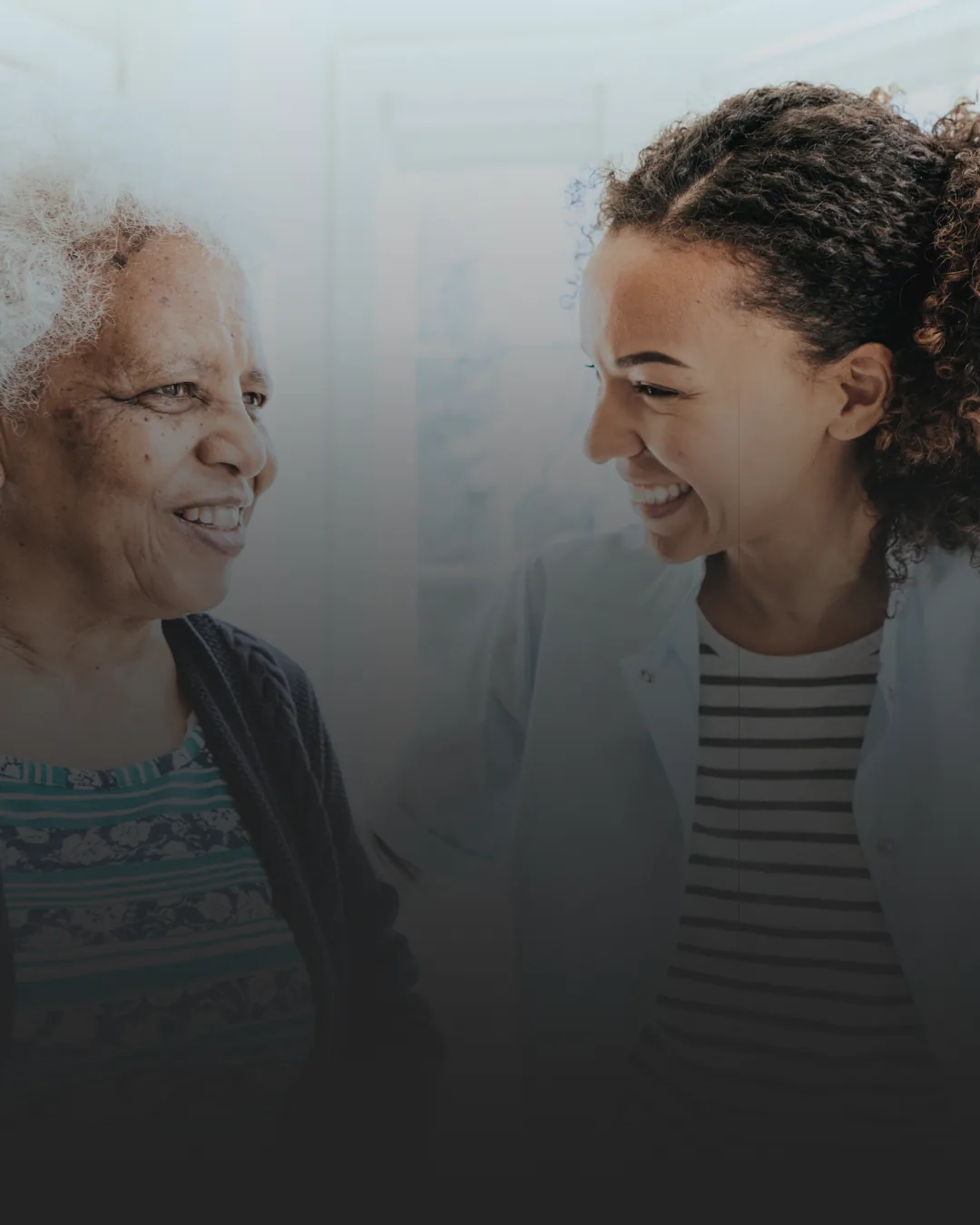 A healthcare professional, smiling, engaging with an elderly woman in a bright room.