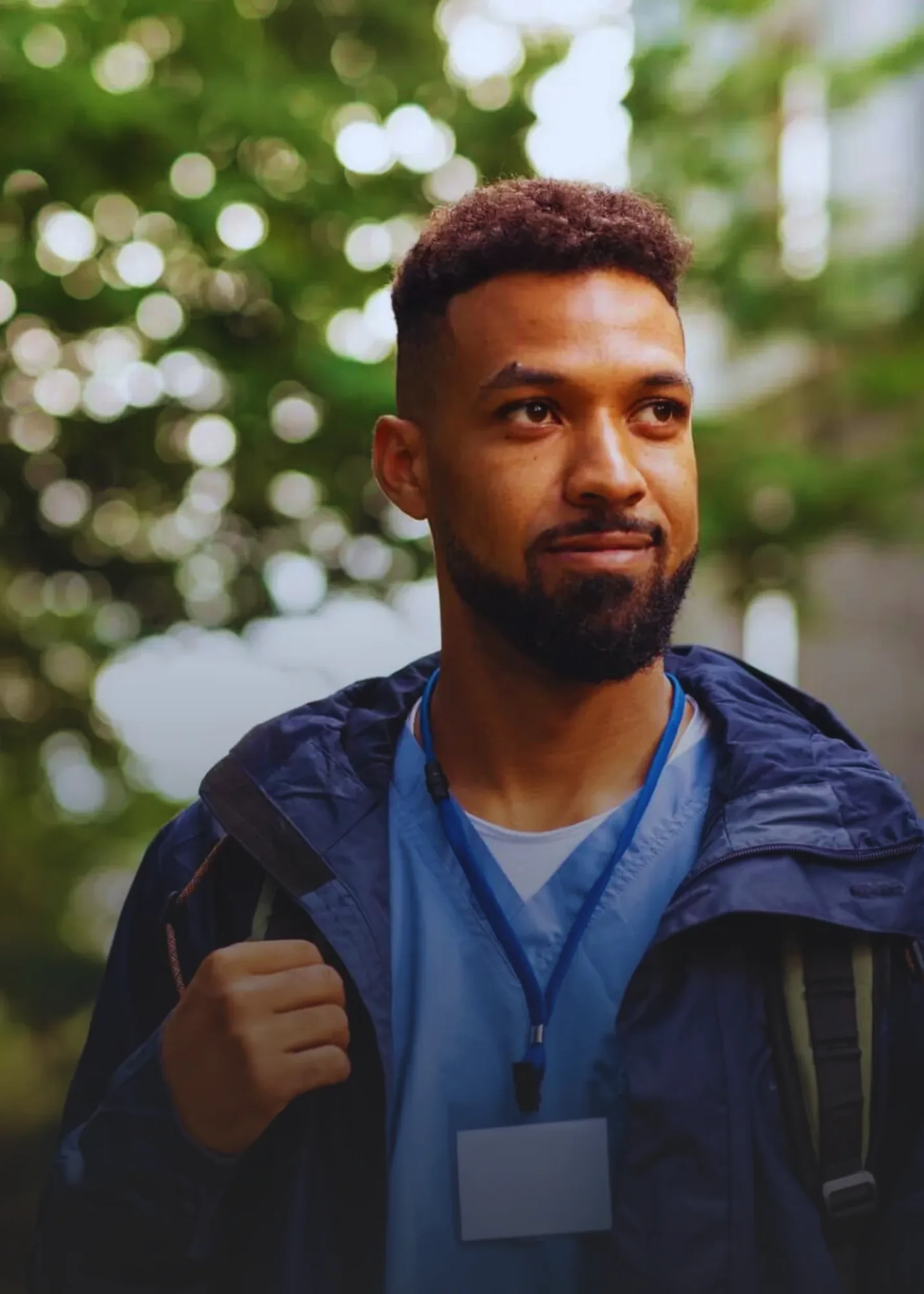 A man outdoors in a natural setting, wearing a blue jacket and a lanyard with a badge, carrying a backpack, and smiling softly.