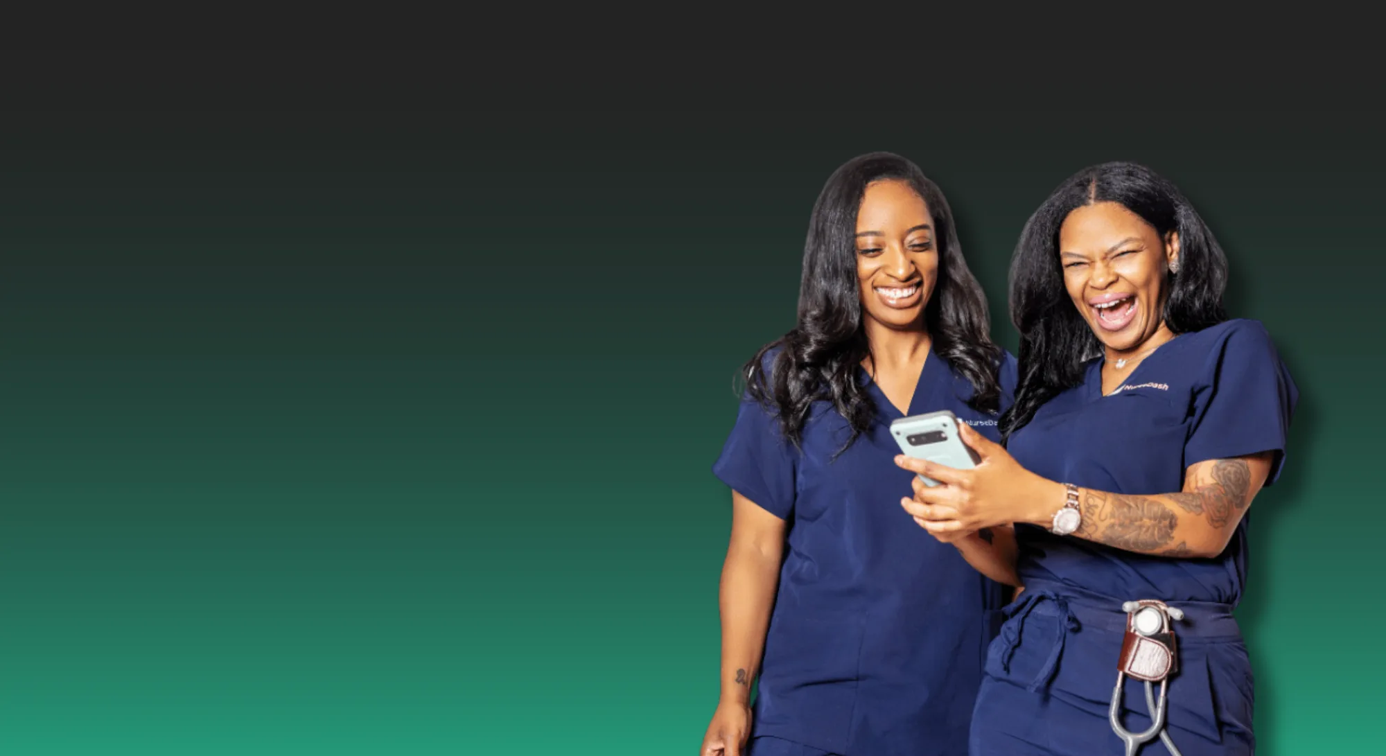 Two women dressed in medical scrubs looking at a smartphone together, one has tattoos on her arm and pocket with medical tools, both smiling and sharing a moment of joy.
